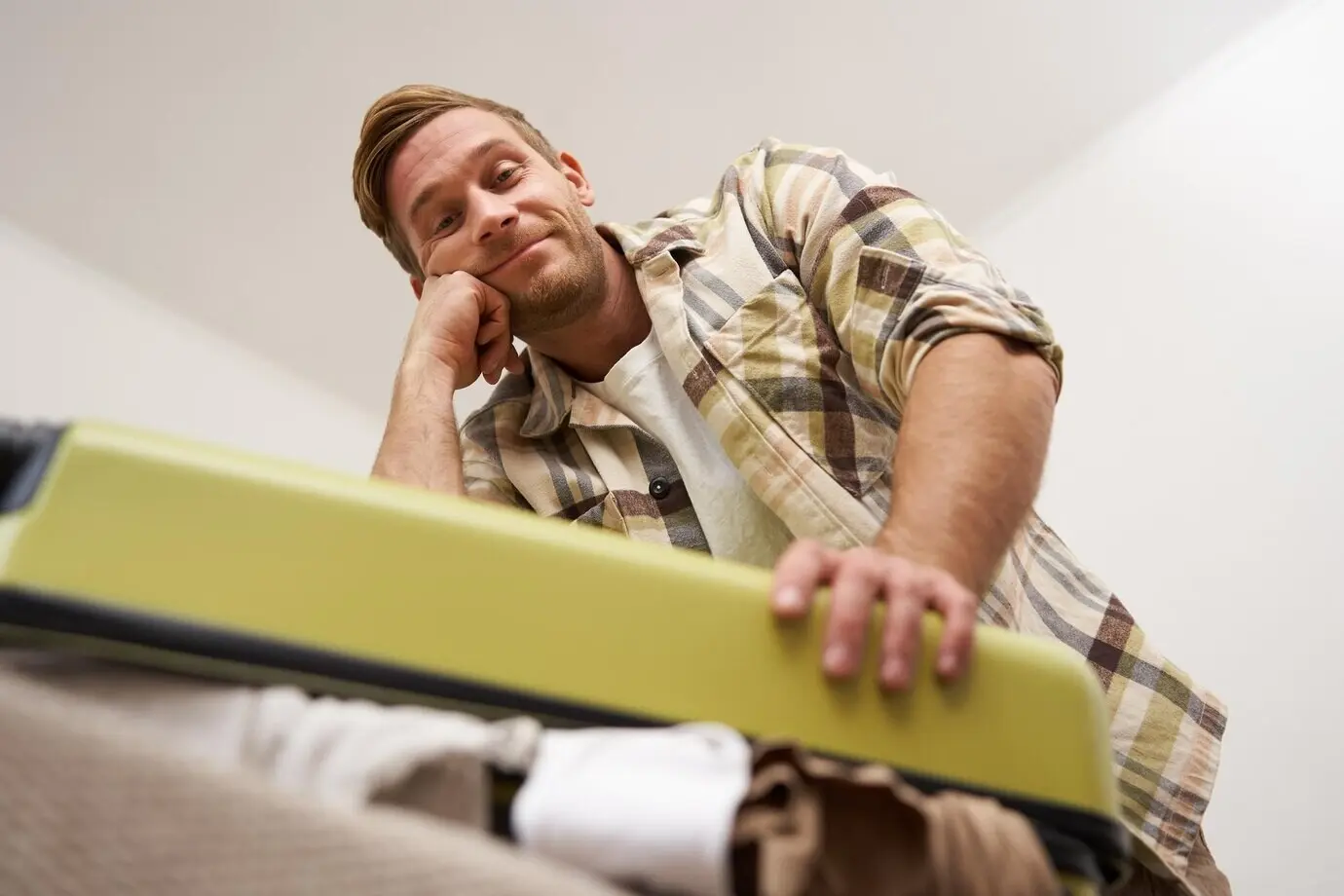An image of a funny man trying to close a suitcase filled with a pile of clothes while packing his luggage.